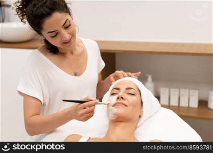 Smiling female cosmetologist applying hydrating mask with brush on face of female customer lying on table in beauty salon. Beautician applying facial mask on client