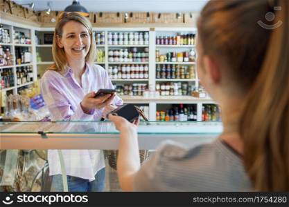Smiling Female Customer Delicatessen Food Store Making Contactless Payment With Mobile Phone For Shopping To Sales Assistant