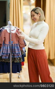 Smiling female customer looking at price of clothes hanging on rail in child store during shopping in mall. Positive female buyer reading label on clothes in child shop