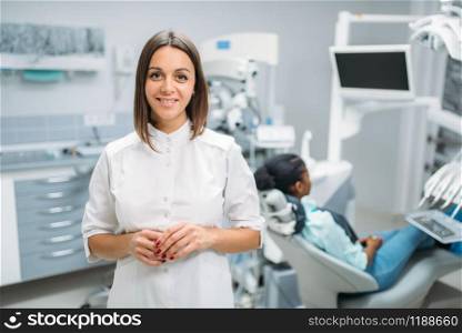 Smiling female dentist, dental clinic, patient in chair on background. Woman in dentistry cabinet, stomatology, teeth care