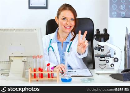 Smiling female doctor sitting at office table and showing victory gesture&#xA;