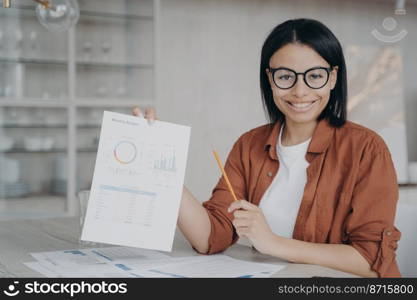 Smiling female financial analyst showing paper with company statistics, presenting project, sitting at desk full of documents. Happy female employee financier shows work report on workplace.. Smiling female financial analyst shows document with company statistics, presents project at desk