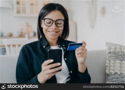 Smiling female holding bank credit card and smartphone. Young woman pays for purchases in online store using banking service, shopping, making secure payment, sitting on sofa at home. E-commerce.. Female holds bank credit card, smartphone, uses online banking services, sitting on sofa at home
