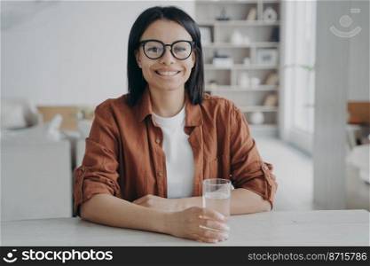 Smiling female holding glass of pure water sitting at table at home. Happy woman wearing glasses holds fresh mineral seltzer, looking at camera. Healthy lifestyle, wellness, diet concept.. Smiling female holding glass of pure water sitting at table at home. Healthy lifestyle, wellness