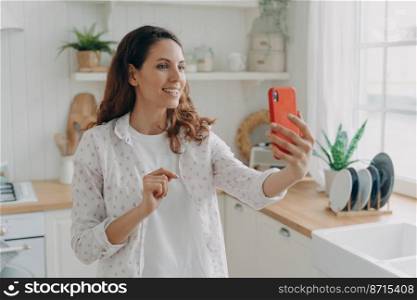 Smiling female holding phone, making video call to friends or relatives, enjoying pleasant conversation in kitchen at home. Happy woman blogger showing her apartment followers online.. Smiling female holding phone, making video call, enjoying online conversation in kitchen at home