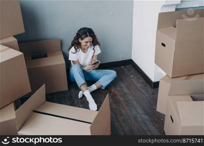 Smiling female holding smartphone, choosing moving company for relocation, sitting on the floor with carton boxes in vacated apartment. Girl selects a reputable mover for removal things.. Modern girl sits with boxes uses smartphone app, choosing moving company and mover for relocation