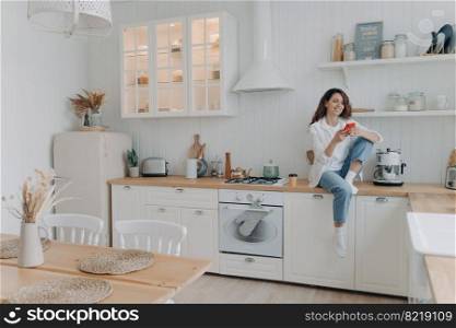 Smiling female homeowner tenant holding phone, makes purchases on internet, sitting in modern kitchen interior. Happy woman housewife using smartphone apps, rests after chores, shopping online at home. Female holding smartphone shopping online by mobile apps, sitting in modern kitchen interior at home