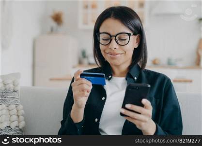 Smiling female in glasses holding bank credit card and smartphone uses online banking service at home. Happy woman pays for purchases, shopping on internet, sitting on sofa. E-commerce.. Female in glasses holds bank credit card, smartphone, uses online banking service at home. E-banking