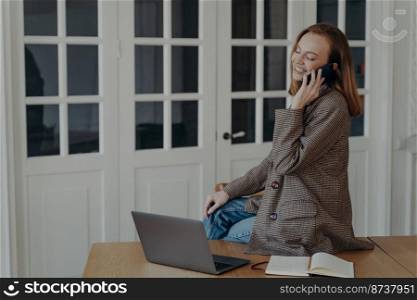 Smiling female manager freelancer answers mobile call, sitting on desk with laptop talks by phone. Happy young woman employee sits at office table chatting, having pleasant cellphone conversation.. Smiling female manager freelancer answers mobile cal, sitting on desk with laptop talks by phone