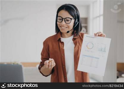Smiling female manager in headset shows document with statistics while online conference at laptop. Friendly businesswoman in headphones having video call, discussing business project.. Smiling female manager in headset shows document with statistics while online conference at laptop