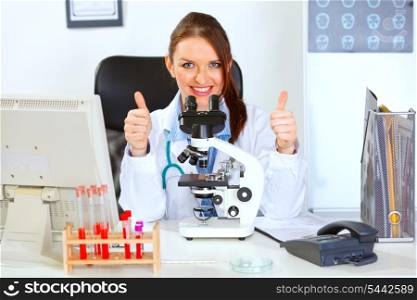 Smiling female medical doctor using microscope in laboratory and showing thumbs up gesture&#xA;