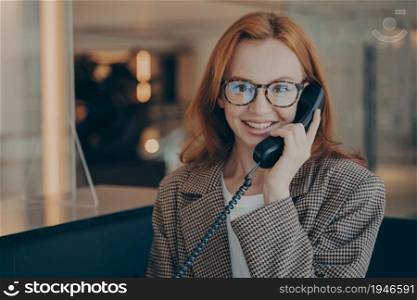 Smiling female office worker with red hair in plaid jacket wearing glasses, talking on landline phone with her co-worker about project completion date, office environment in blurred background. Female office worker in spectacles talking on landline phone with co-worker and smiling at camera
