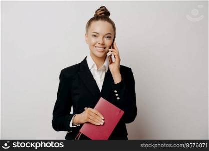 Smiling female office worker, woman in formal black costume having telephone conversation, holding red note book with data, hearing good news while standing isolated on grey background. Happy businesswoman talking on phone and smiling at camera