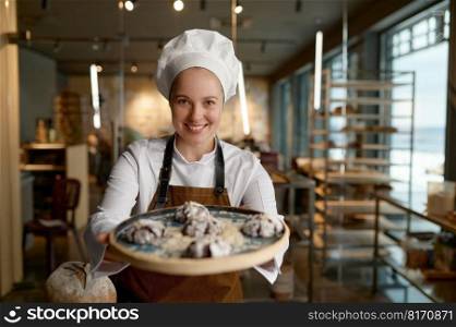 Smiling female pastry chef presenting freshly baked cookies standing at bakery kitchen. Happy confectioner showing delicious freshly made sweet brownie on tray. Pastry chef presenting freshly baked cookies at bakery kitchen