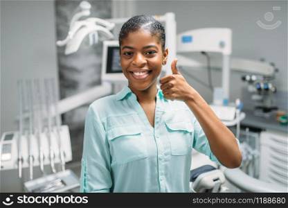 Smiling female patient shows thumbs up in dental clinic, medical equipment on background. Woman in dentistry cabinet, stomatology, teeth care. Female patient shows thumbs up in dental clinic