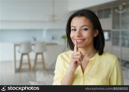 Smiling female puts finger on lips, makes hush gesture at home. Happy woman share secret private information, ask silence, looking at camera. Unique sale offer, big discounts in online store.. Female makes silence gesture, share secret at home. Unique sale offer, big discounts in online store