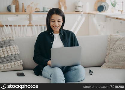 Smiling female shopping on the internet at laptop sitting on couch at home. Happy woman freelancer working on computer, chatting in social networks. Ecommerce, online store advertisement.. Smiling female shopping on the internet at laptop sitting on couch at home. Ecommerce, online store