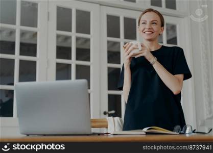 Smiling female standing near workplace desk with laptop starts workday with morning coffee mug. Pensive dreamy young caucasian woman looks in distance lost in thoughts, pondering, visualizing.. Smiling female standing near workplace desk with laptop starts workday with morning coffee mug