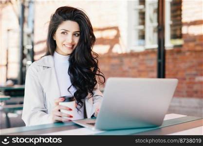 Smiling female student with dark wavy hair reading news on her laptop having pleased expression while looking aside resting in cafe after classes drinking takeaway coffee admiring fresh air outdoors