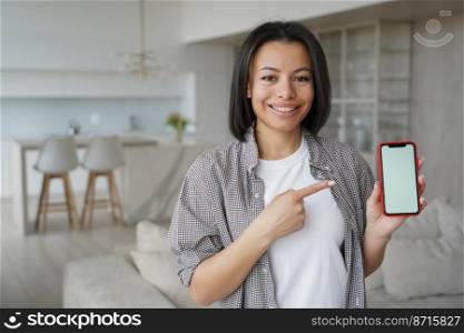 Smiling female tenant pointing at smartphone with mockup blank screen at home. Happy woman renter showing template phone screen, recommends house hunting app, service for housing search.. Female tenant pointing at smartphone with mockup blank screen recommends house hunting app at home