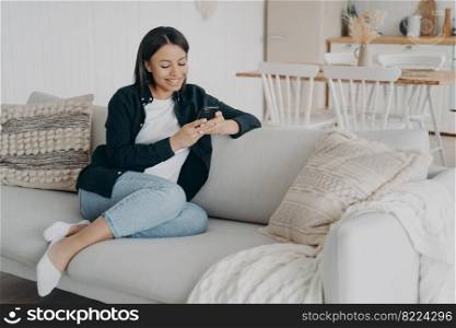 Smiling female uses phone apps, chatting in social networks, shopping online, sitting on couch at home. Happy young woman holding smartphone, enjoying internet services, browsing good news. E-commerce. Smiling female uses phone apps, chatting in social networks, shopping online, sits on couch at home