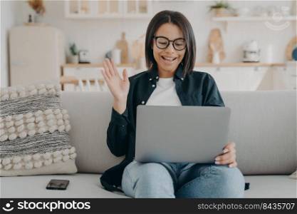 Smiling female waving hand to laptop greeting teacher at lesson consultation, sitting on couch at home. Friendly young woman say hello to friend speaking by video call. Online communication, elearning. Smiling female waving hand to laptop greeting teacher at lesson, sitting on couch at home. Elearning