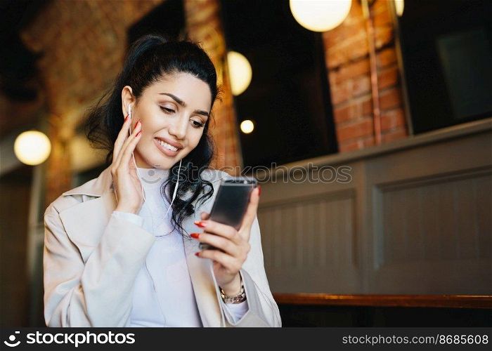 Smiling female with dark hair tied in pony tail wearing white clothes holding smartphone while listening to music on her electronic device. Stylish woman resting in cafe using app having smile on face