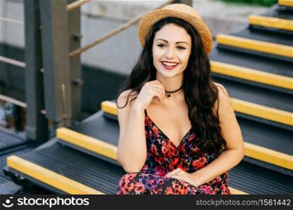 Smiling female with dark luxurious hair wearing dress and straw hat sitting at stairs looking with delightful look into distance. Restful young woman with appealing appearance sitting outdoors