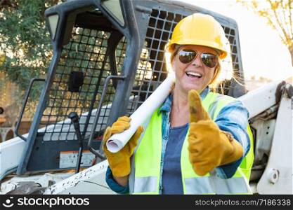 Smiling Female Worker With Thumbs Up and Technical Blueprints Near Small Bulldozer At Construction Site