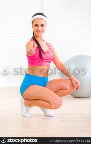 Smiling fitness young girl squatting down and showing thumbs up gesture&#xA;