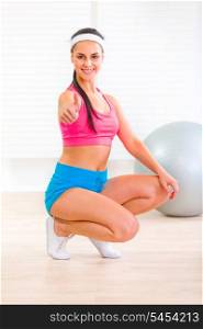 Smiling fitness young girl squatting down and showing thumbs up gesture&#xA;