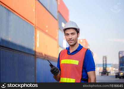 Smiling foreman in hardhat and safety vest talks on two-way radio control loading containers box from cargo