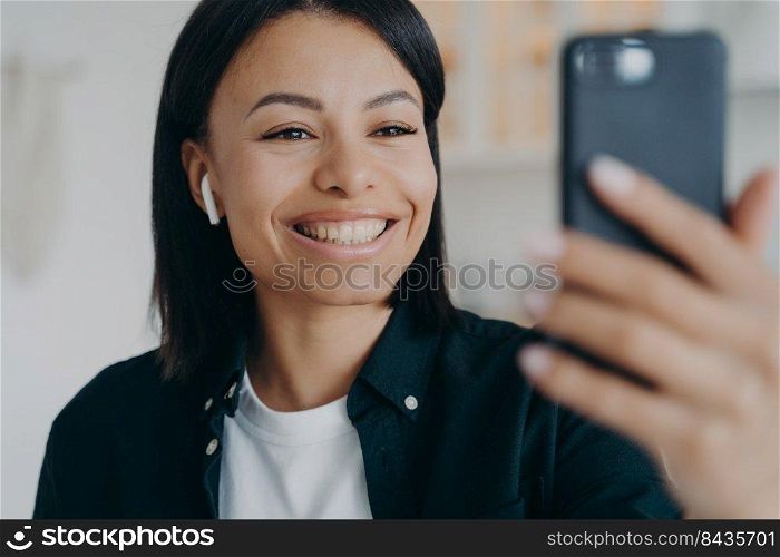 Smiling friendly young woman wearing earphone communicate on video call holding smartphone in hand. Happy female in earphones having online communication on cellphone, using webcam.. Smiling friendly young woman wearing earphone communicate on video call holding smartphone in hand
