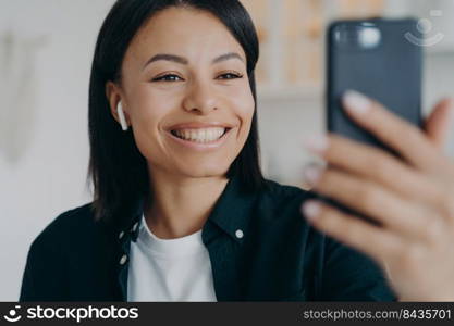 Smiling friendly young woman wearing earphone communicate on video call holding smartphone in hand. Happy female in earphones having online communication on cellphone, using webcam.. Smiling friendly young woman wearing earphone communicate on video call holding smartphone in hand