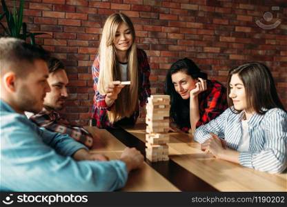 Smiling friends plays table game at home, selective focus on tower. Board game with wooden blocks requiring high concentration, entertainment for funny company. Friends plays table game, selective focus on tower