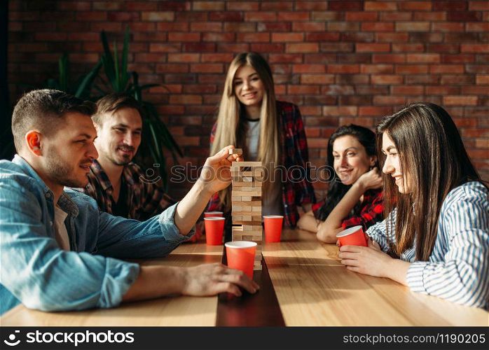 Smiling friends plays table game at home, selective focus on tower. Board game with wooden blocks requiring high concentration, entertainment for funny company. Friends plays table game, selective focus on tower