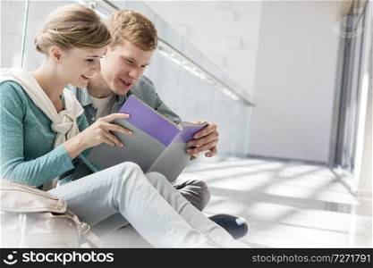 Smiling friends reading book while sitting on corridor at university campus