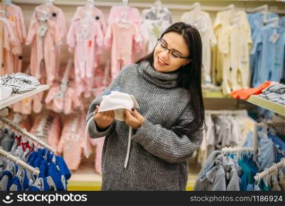 Smiling future mother chooses baby hat in the store for newborns. Pregnant woman in shop of goods for infants. Smiling future mother chooses baby hat