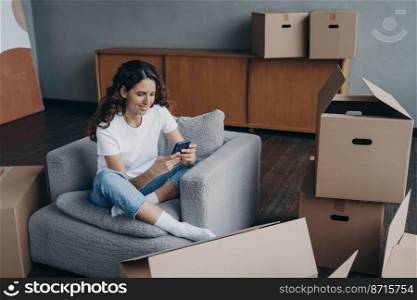 Smiling girl choosing mover online, using mobile phone app, sitting with cardboard boxes waiting to move to new home. Satisfied woman holding smartphone, arranging relocation, selects moving company.. Smiling girl holding smartphone, selects moving company online for relocation, sitting with boxes