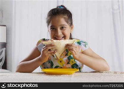 Smiling girl eating a bread sandwich sitting at dining table