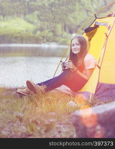 smiling girl hiker in a tent and holding a cup mountains in the background