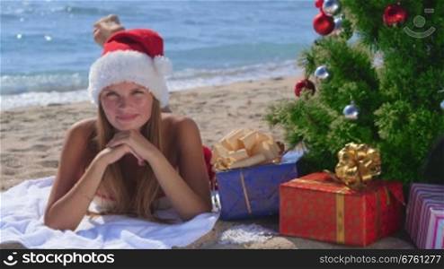 Smiling girl in Santa hat with gift boxes under Christmas tree on a sandy beach looking at camera