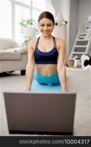 Smiling girl sits on the floor at home, online fit training at the laptop. Female person in sportswear, internet sport workout, room interior. Smiling girl sits on floor, online fit training