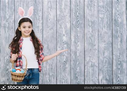 smiling girl with bunny ears holding basket easter eggs presenting against gray wooden desk