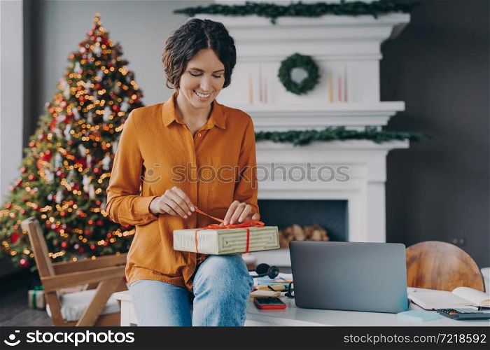 Smiling glad italian female tying red ribbon on Xmas present while sitting at home office with Christmas tree on background, woman preparing gifts for family and friends during winter holidays. Smiling glad italian female tying red ribbon on Christmas present while sitting at home office