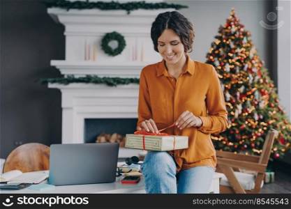 Smiling glad italian female tying red ribbon on Xmas present while sitting at home office with Christmas tree on background, woman preparing gifts for family and friends during winter holidays. Smiling glad italian female tying red ribbon on Christmas present while sitting at home office