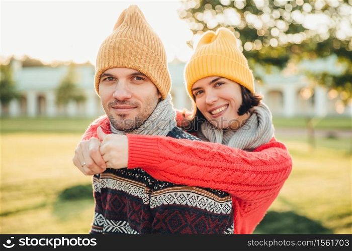 Smiling glad young female in hat and warm cotton sweater embraces her husband who stands back, enjoy spending weekends together, have walk in garden or park, admire splendid sunny autumn weather