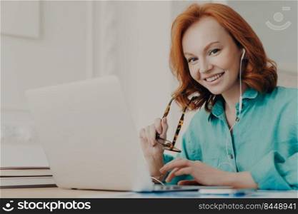 Smiling good looking female employee talks on video call, has web conference with colleagues, watches webinar on modern laptop, poses in coworking space. Student learns foreign languages online