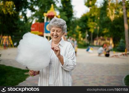 Smiling granny holds cotton candy in summer amusement park. Aged people lifestyle. Funny grandmother having fun outdoors, old female person on nature. Smiling granny holds cotton candy, amusement park