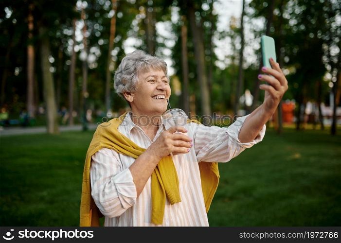 Smiling granny makes selfie with milk coctail in summer amusement park, back to childhood. Aged people lifestyle. Funny grandmother having fun outdoors, old female person on nature. Smiling granny makes selfie with milk coctail
